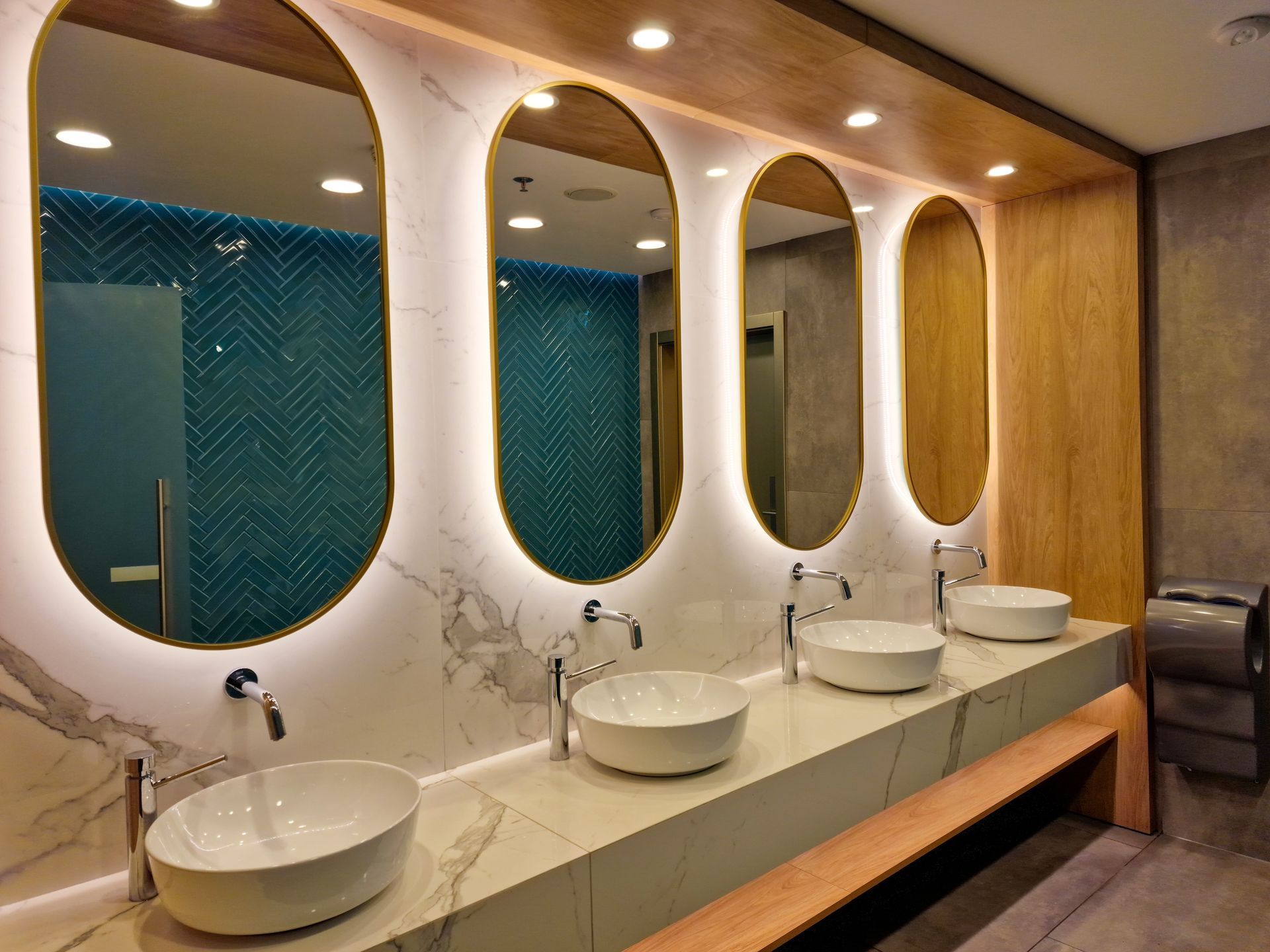 Modern restroom vanity with four oval backlit mirrors above white vessel sinks on a marble countertop.