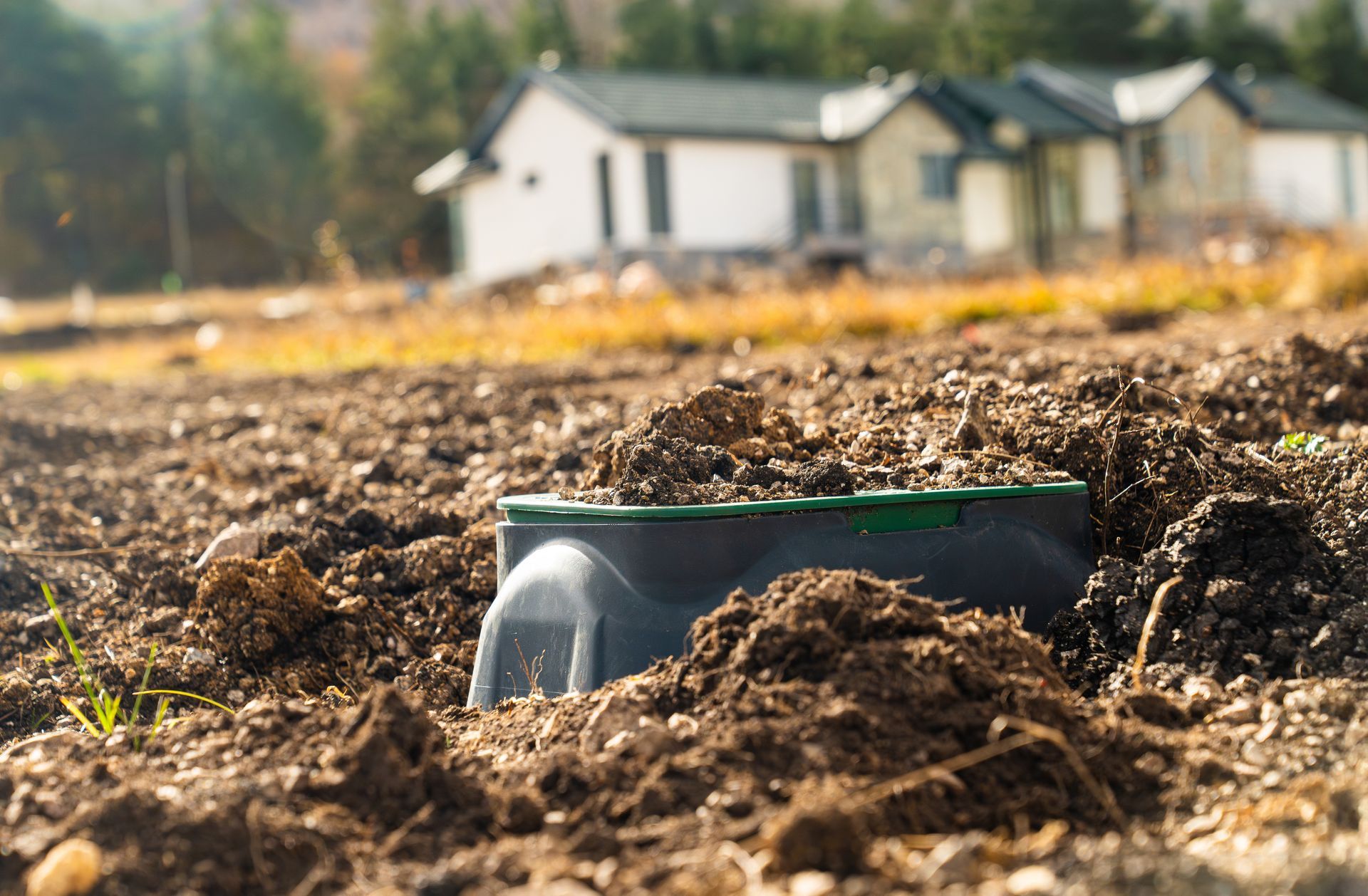 A green-lidded utility box sits partially buried in soil with a house blurred in the background.