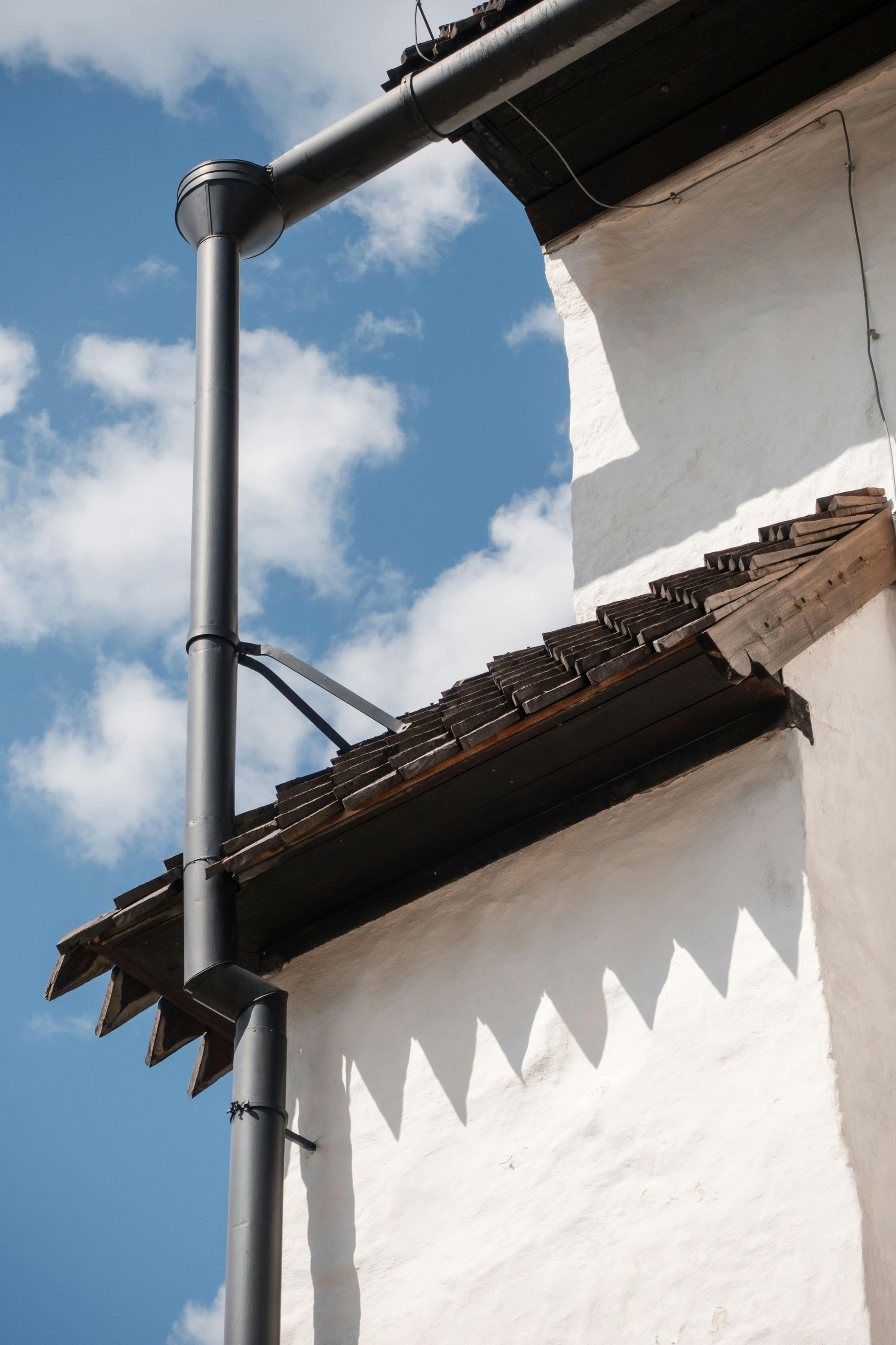 White building corner with a black downspout and rusted roof edge against a blue sky