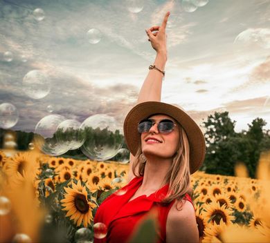 A woman wearing sunglasses and a hat is standing in a field of sunflowers.