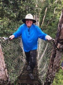 A man in a blue shirt and hat is walking across a rope bridge.