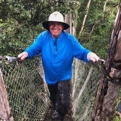 A man in a blue shirt and hat is walking across a rope bridge.