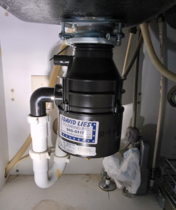 Under-sink garbage disposal and plumbing with black unit and white PVC pipes.