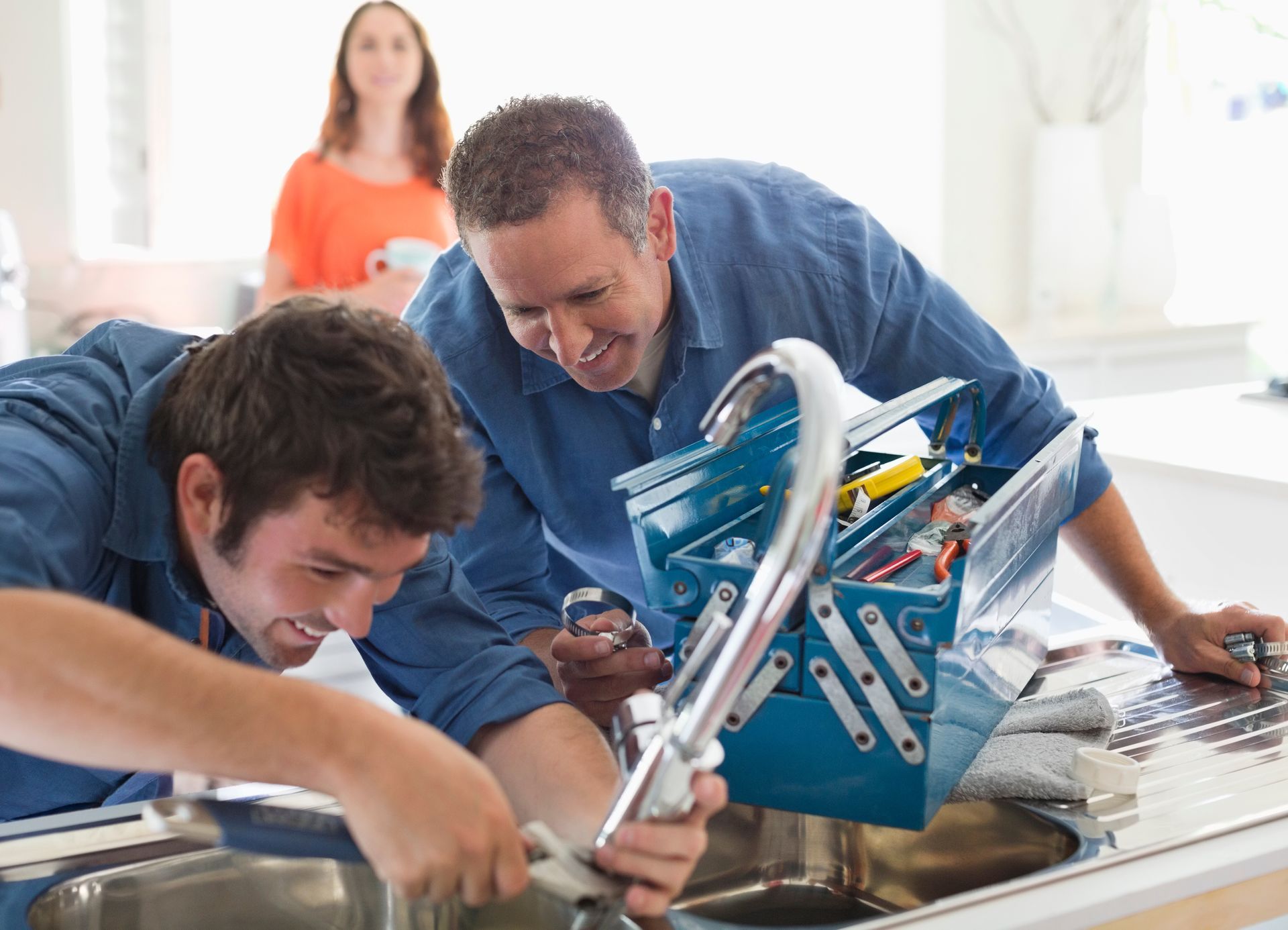 Two plumbers fixing a kitchen sink faucet with a tool box and a customer in the background.