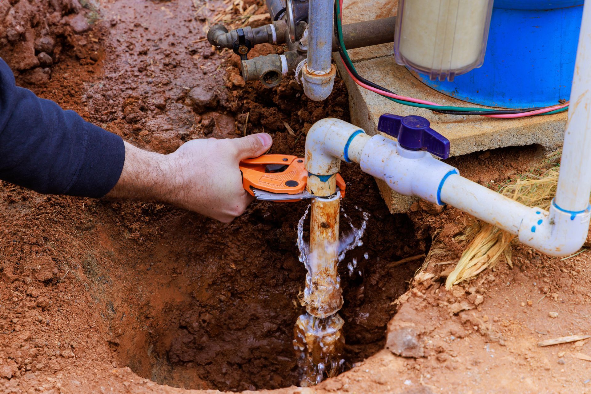 Hand digging around a buried water pipe with white PVC and blue valve fittings.