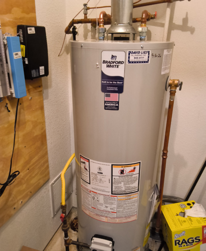 A plumber is adjusting a shower head, using tools to ensure proper installation and functionality. Water heater in a utility closet with pipes, wall controls, and a yellow rag bag nearby.