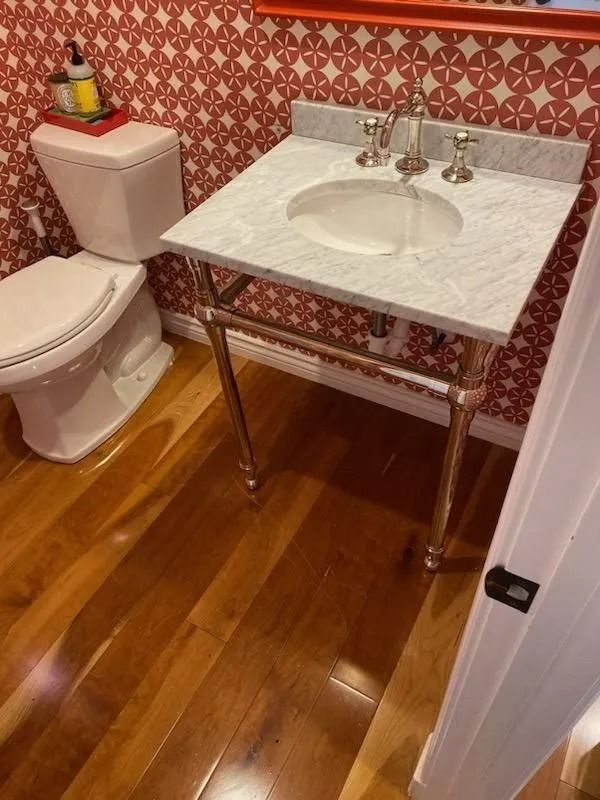 Small bathroom with toilet, marble sink vanity, red patterned wallpaper, and wood floor