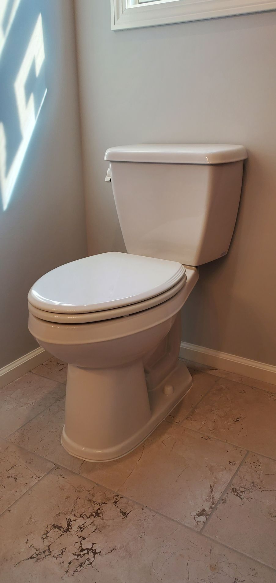 White toilet in a beige bathroom corner with tiled floor and a window casting light on the wall