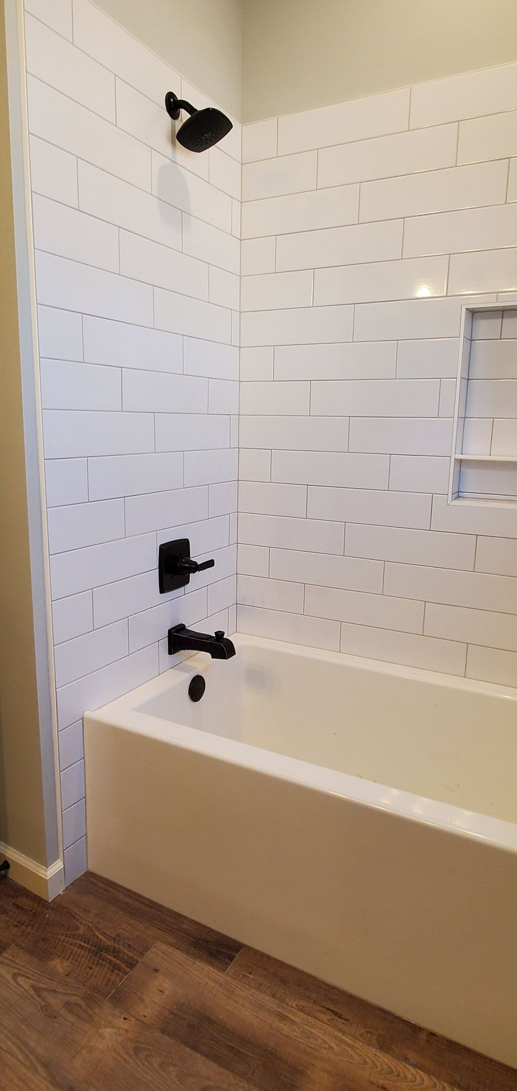 White-tiled bathtub and shower with black fixtures and wood floor in a bathroom corner
