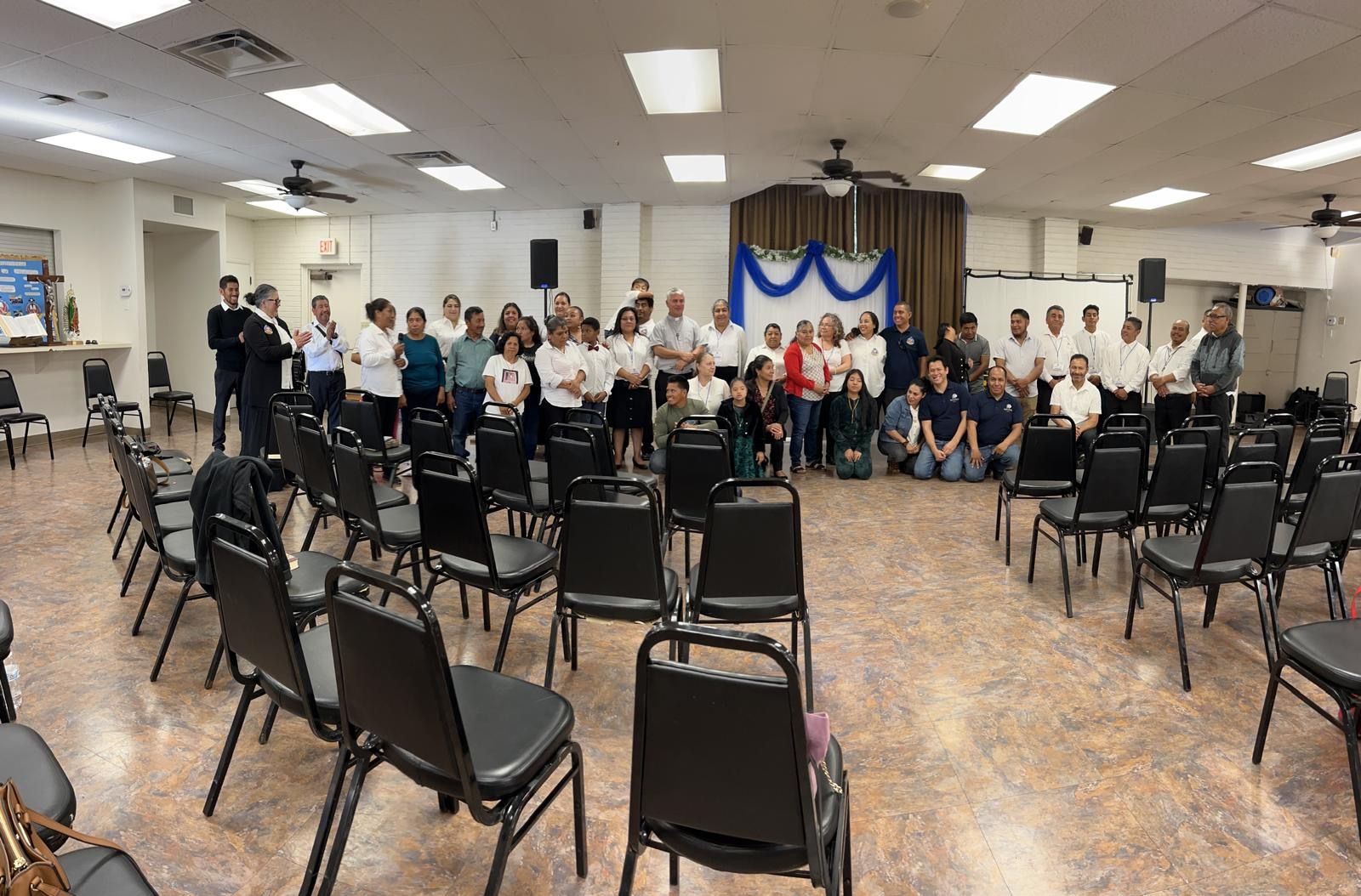 Community event in a hall, with chairs arranged facing a decorated stage and a group gathered at the front.