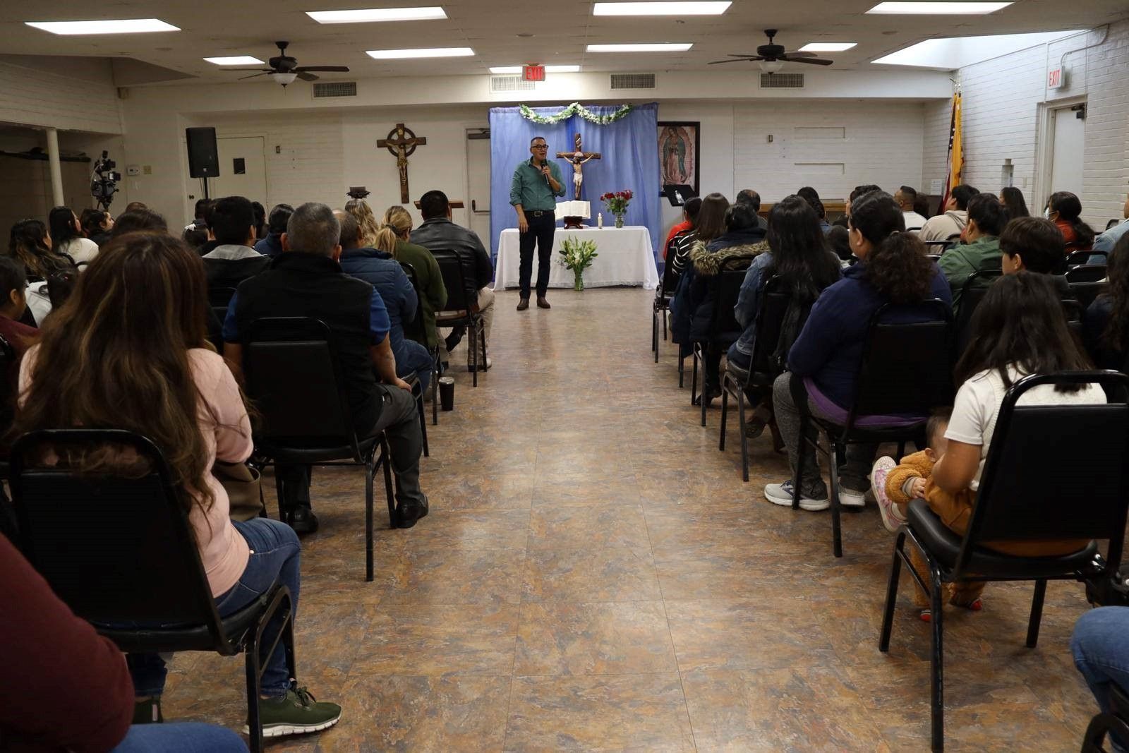Audience seated in a hall facing a speaker at a podium with blue drapes and potted plants.