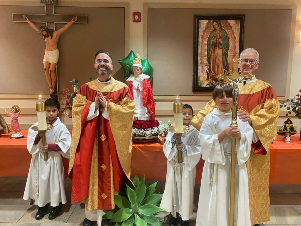 Clergy and altar servers in red and gold vestments stand before a church altar with a crucifix and statue.