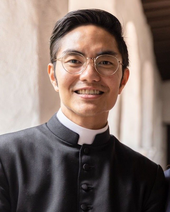Smiling person in a black clerical robe and glasses, standing in a softly lit corridor.