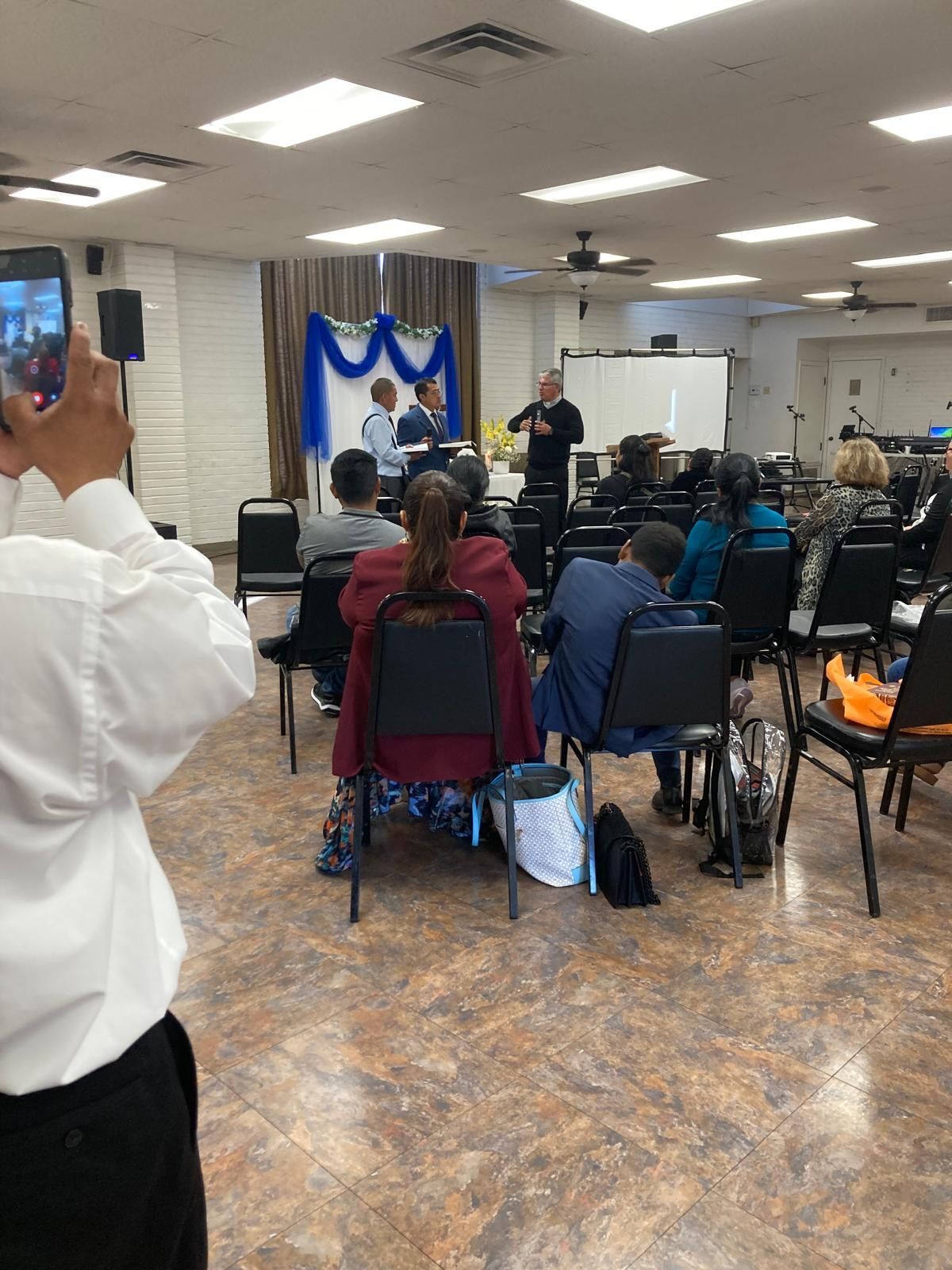 Audience seated in a community hall watching a speaker on stage with blue-and-white drapes.