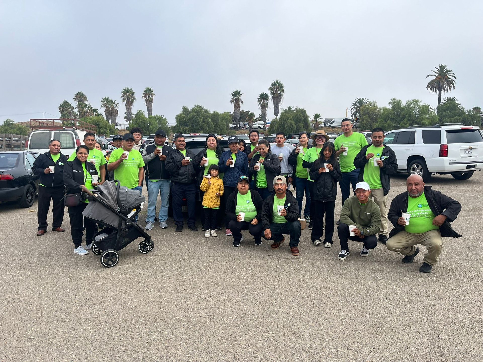 Group of people in neon safety vests posing outdoors in a parking lot with cars and palm trees.