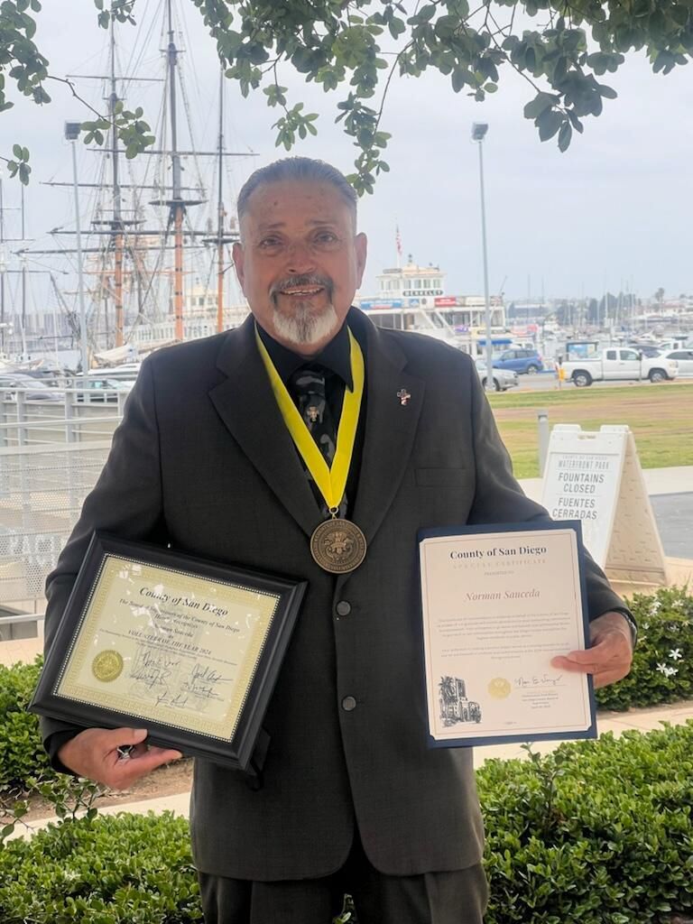 Man in a suit holds a plaque and certificate at a marina, wearing a medal and smiling.