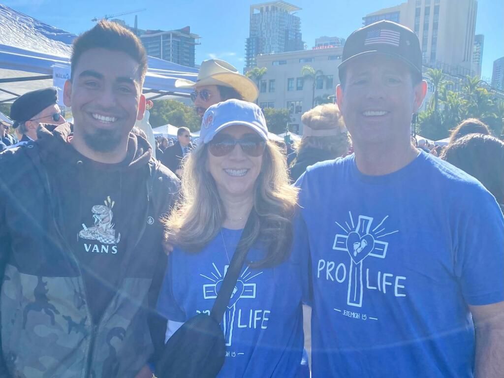 Three smiling people in blue shirts standing outdoors at a sunny event, with buildings in the background