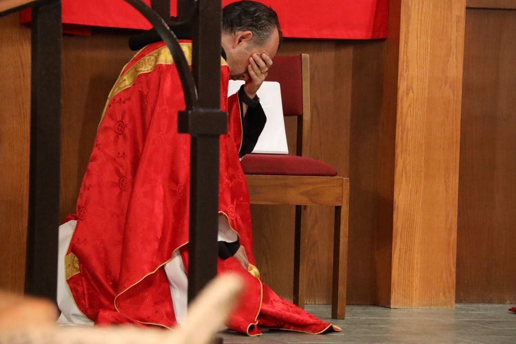 Person in a red robe kneeling beside a wooden chair in a church-like setting
