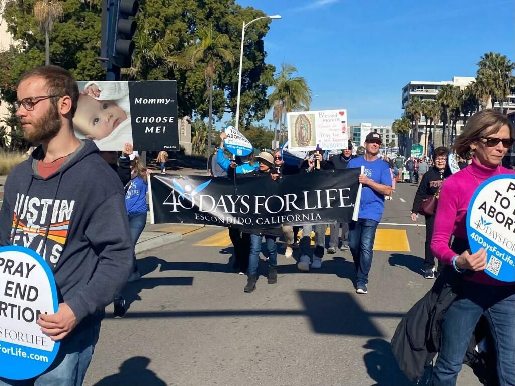 Protesters march on a sunny street carrying signs, including “A 1000 Laws for Life” and “Pray to End Abortion.”