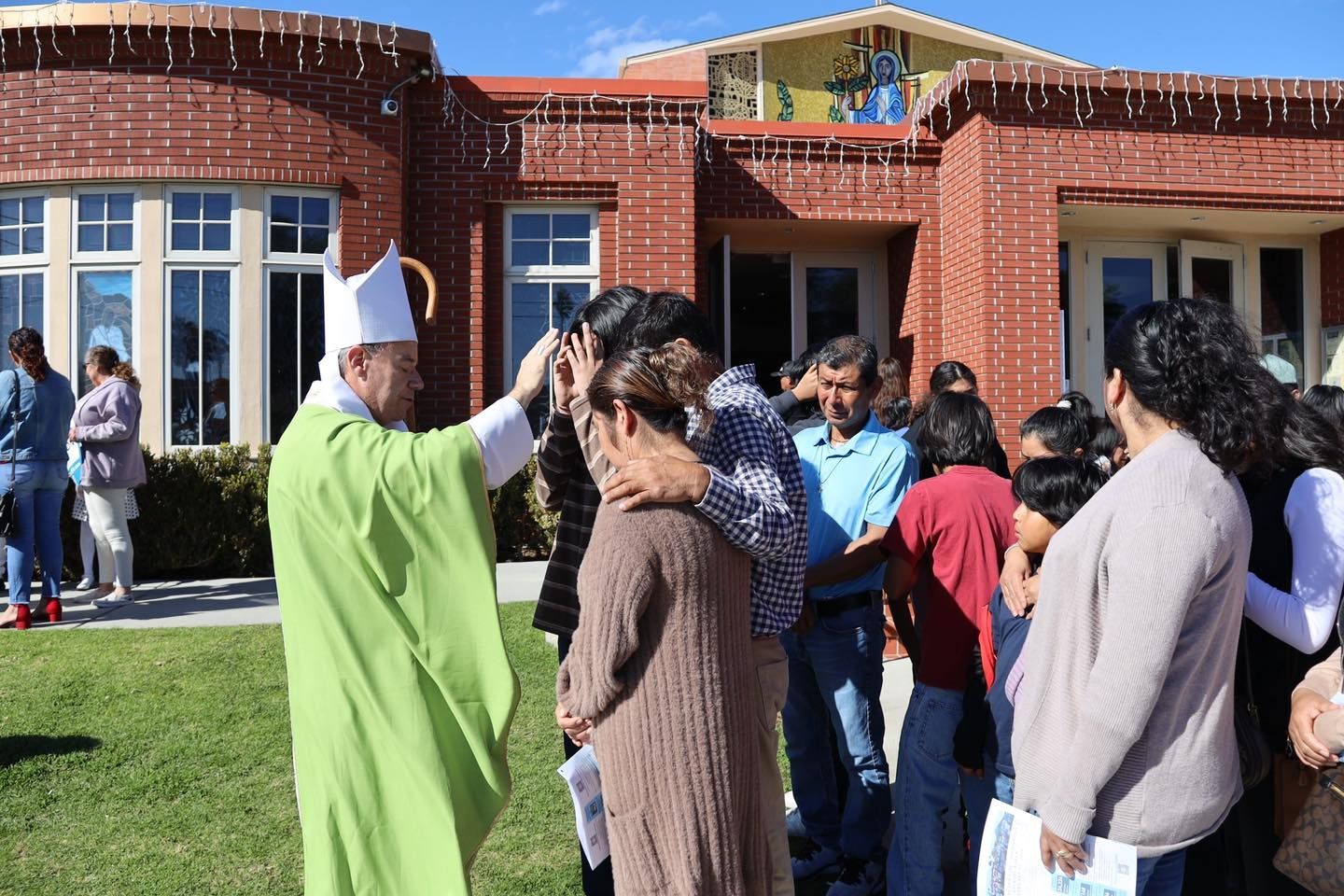 People gathered outside a brick building as a man in a green robe speaks to a small crowd.