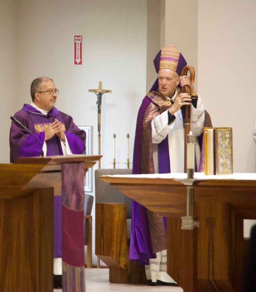 Two clergy in purple vestments at church altar, one reading and one holding a crosier.