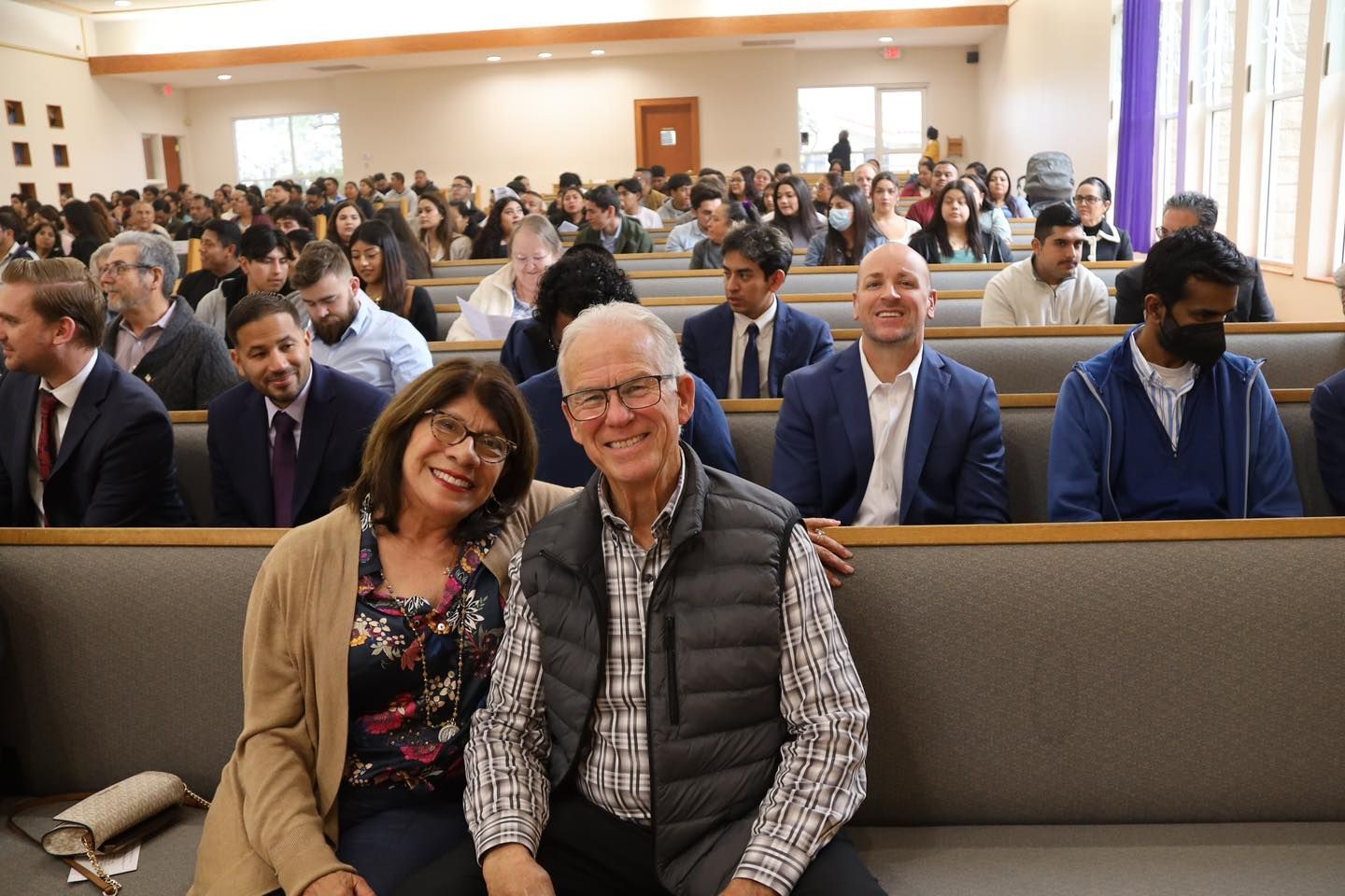 Audience seated in a bright auditorium, with two people smiling in the front row.