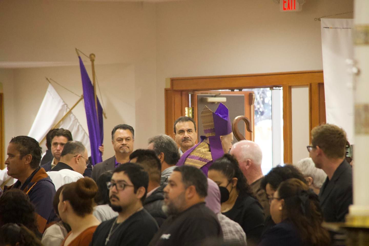 Crowd gathered indoors under purple ribbons near a doorway, facing forward in a hall