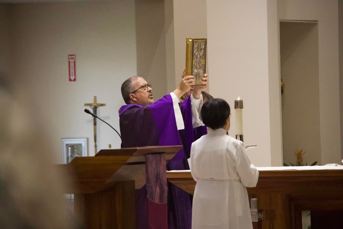 Priest in purple vestments elevates the Eucharist during a church service, with an altar server nearby.