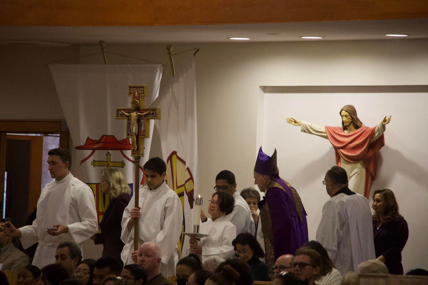 Clergy in white and purple robes process inside a church beside a statue of Jesus with outstretched arms.