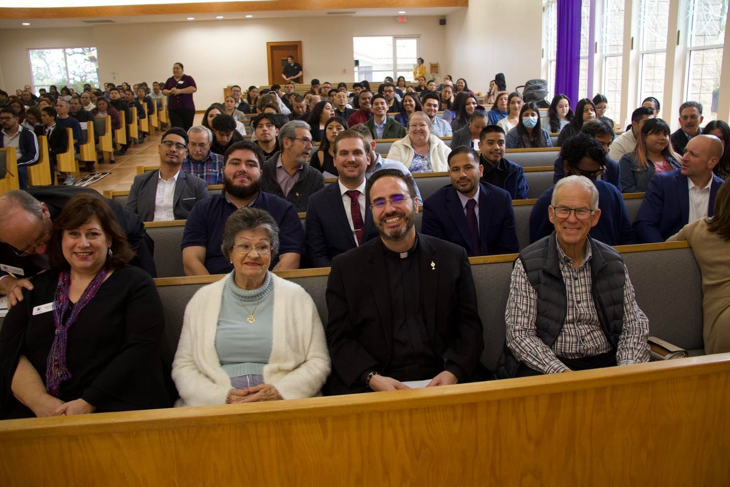 Audience seated in a church or auditorium, facing the camera with rows of people behind them.