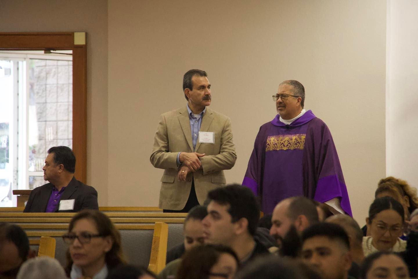 Two men standing and talking in a meeting room, one in a tan suit and one in a purple robe