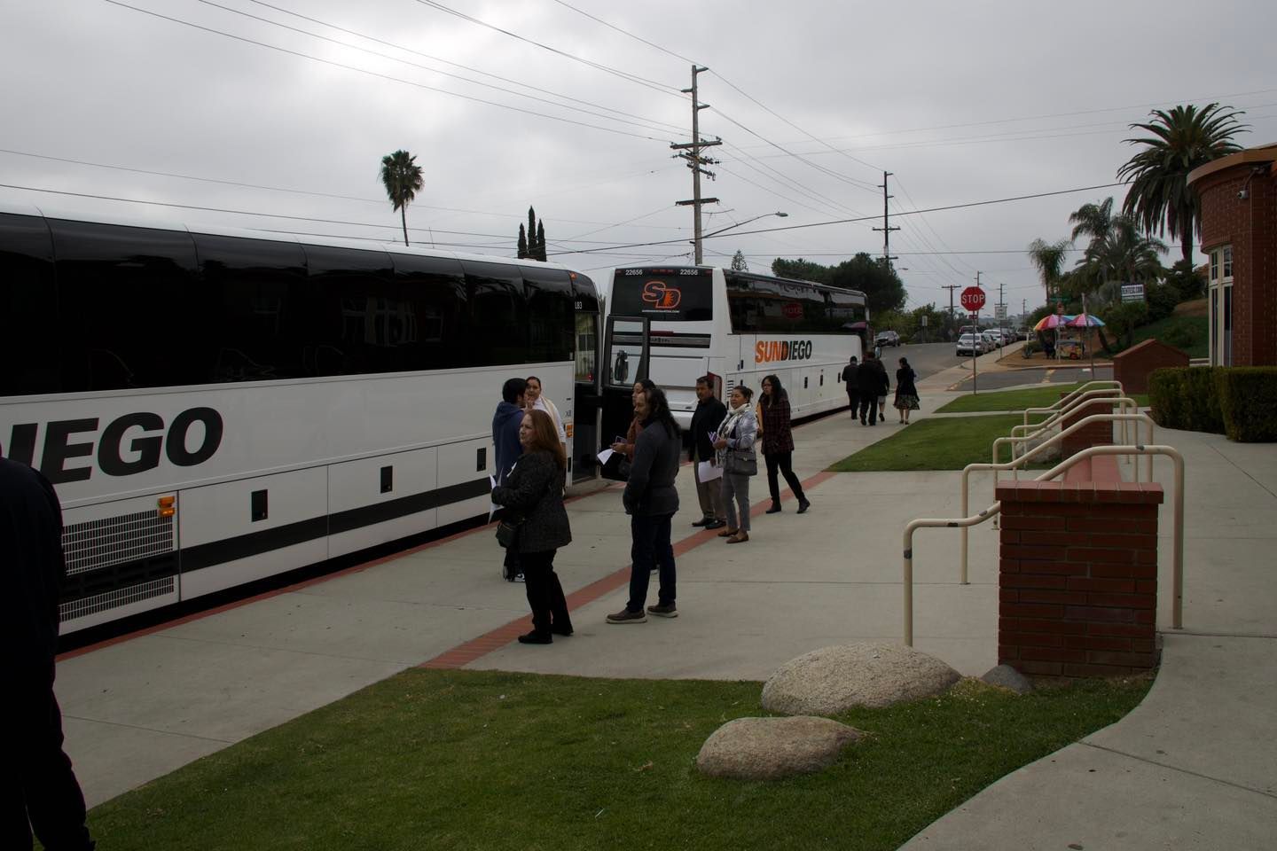 People boarding white coach buses on a suburban street near a sidewalk and lawn under cloudy skies