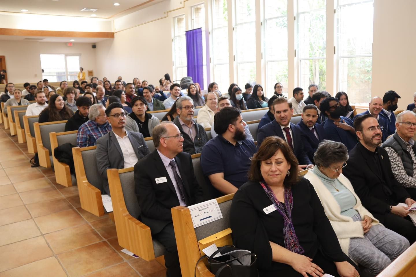 Audience seated in a bright lecture hall, facing forward during a presentation.