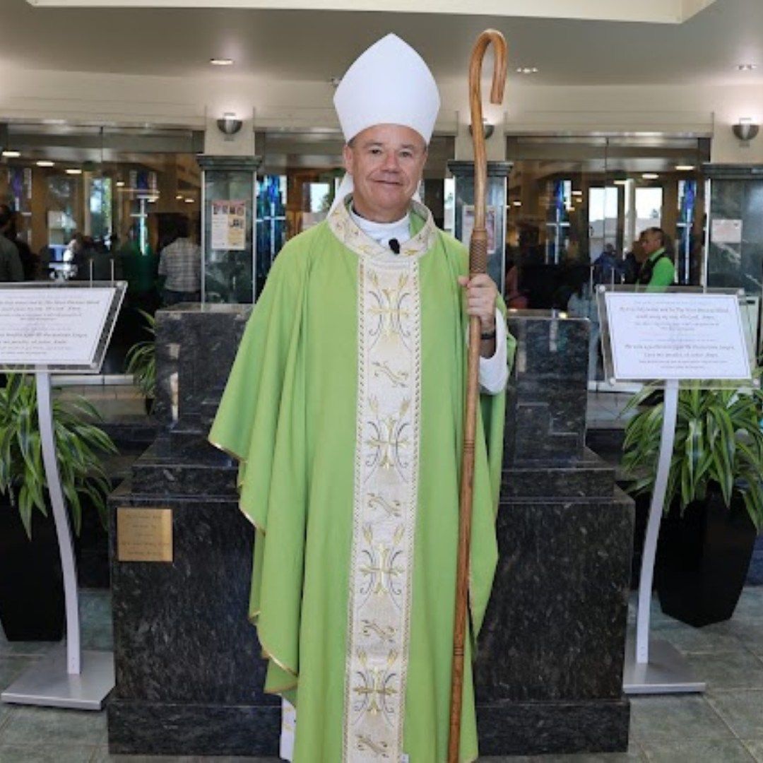 Person in green and white liturgical vestments with a bishop’s staff, standing indoors in a lobby.