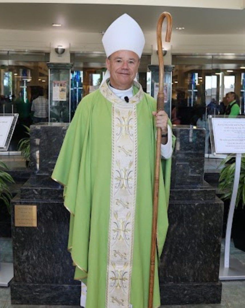 Clergy member in green vestments and white mitre holding a staff indoors.