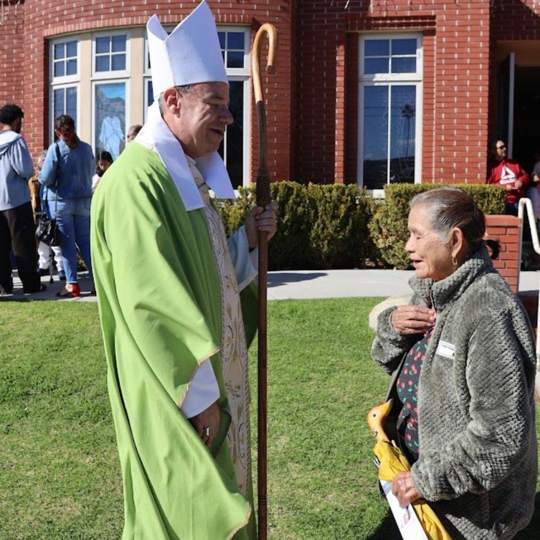 Clergy in green robes greets an elderly woman outside a brick building on a sunny day