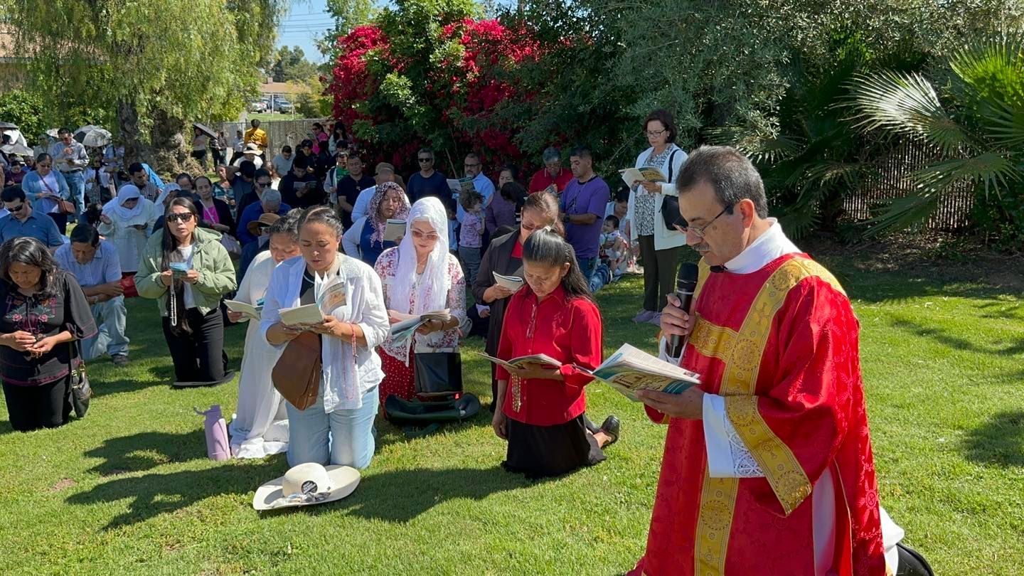 People gathered outdoors for a prayer service, with a priest in red vestments leading on a grassy lawn.