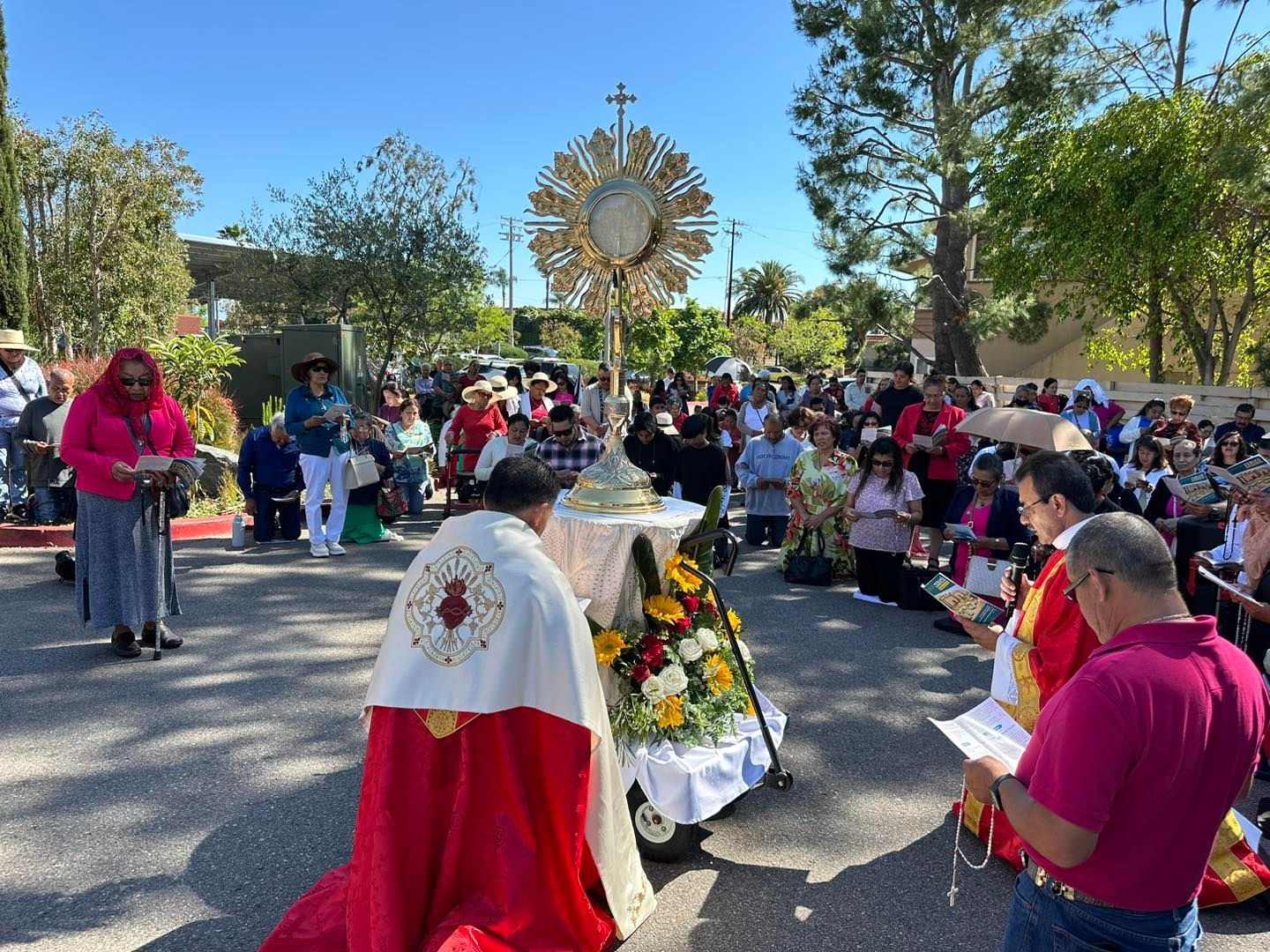Outdoor religious procession with priest in red robes, crowd, and golden monstrance on a sunny street