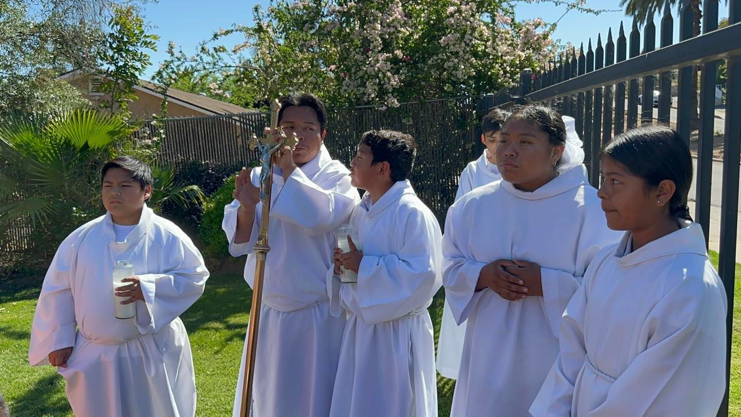 Five people in white robes standing outdoors by a fence in bright sunlight.