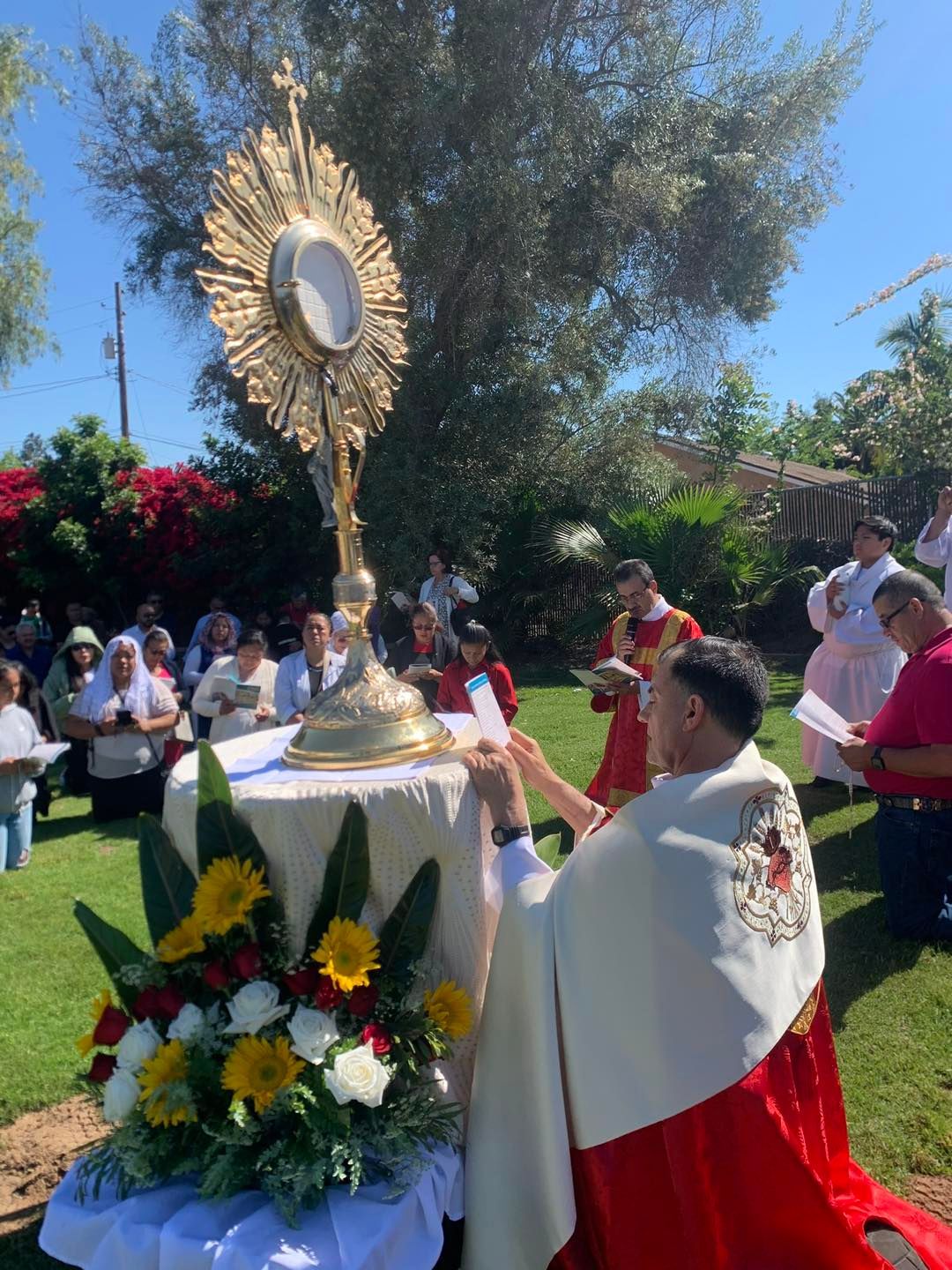 Catholic procession outdoors, with a priest in red and white facing a sunburst monstrance on an altar with flowers