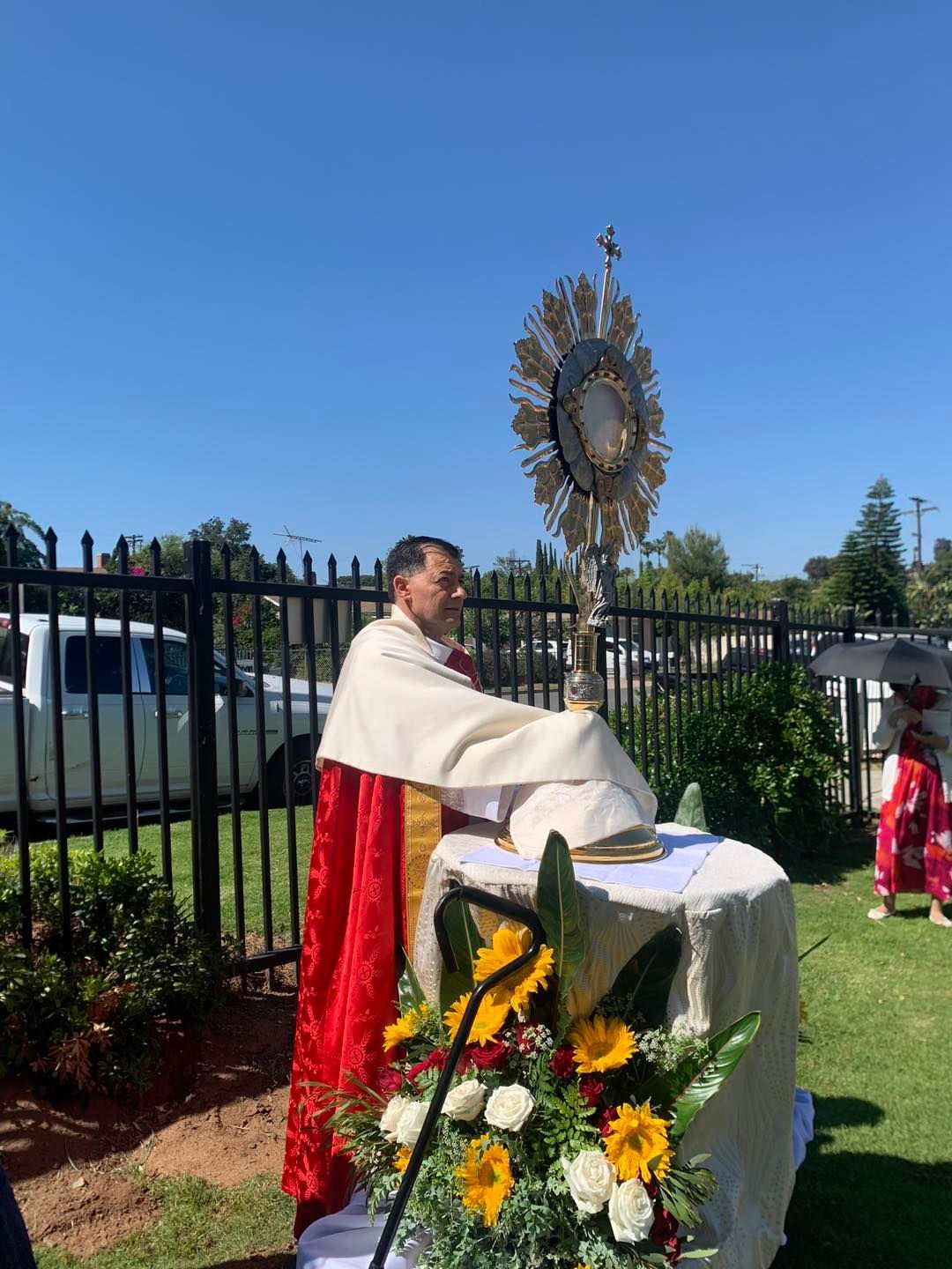 Priest at outdoor altar during Catholic ceremony, with flowers and a monstrance on a sunny lawn.