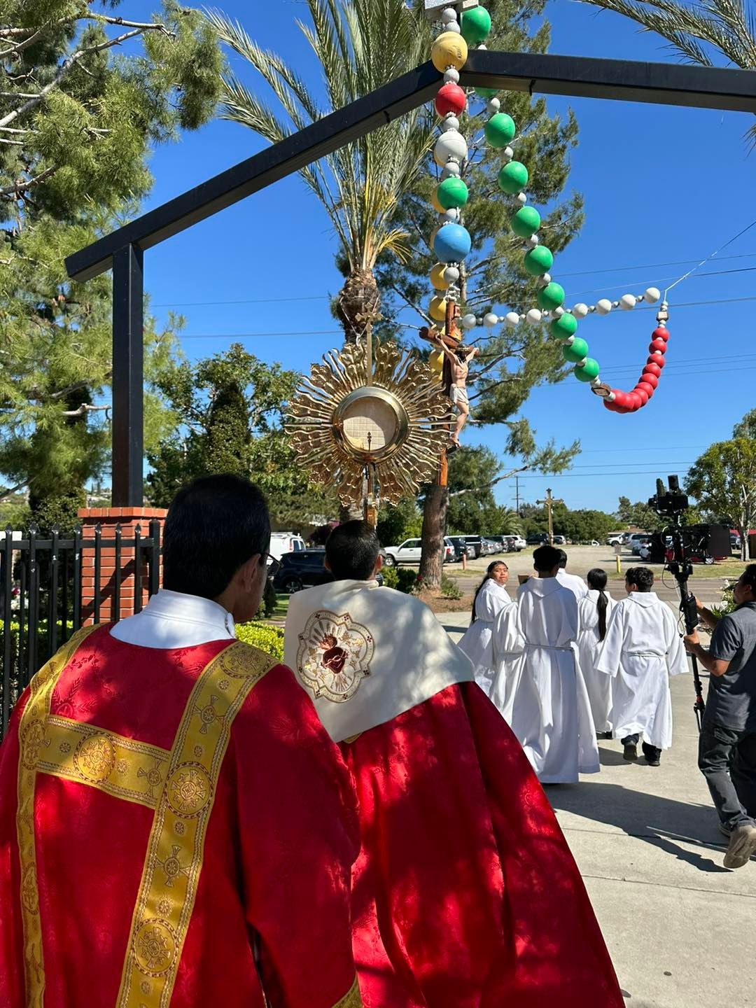 Clergy in red and white robes walking outdoors beneath festive hanging ornaments and a large archway