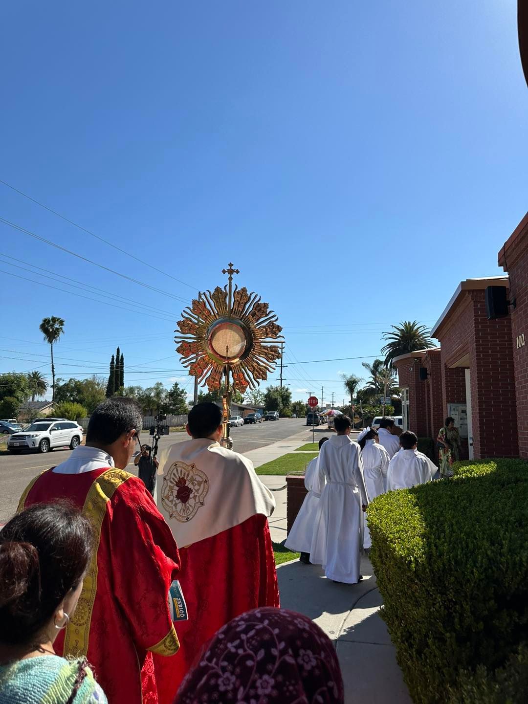 Procession in red and white robes walking toward a sunburst religious display outdoors on a sunny day