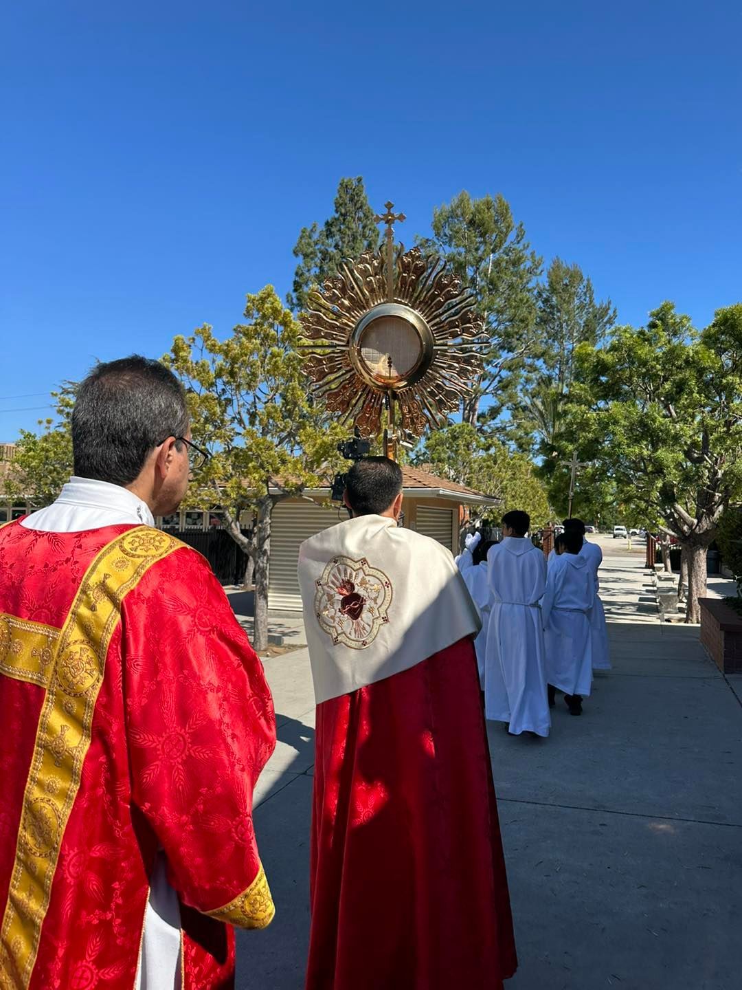 People in red and white robes walking in a sunlit procession beneath a blue sky