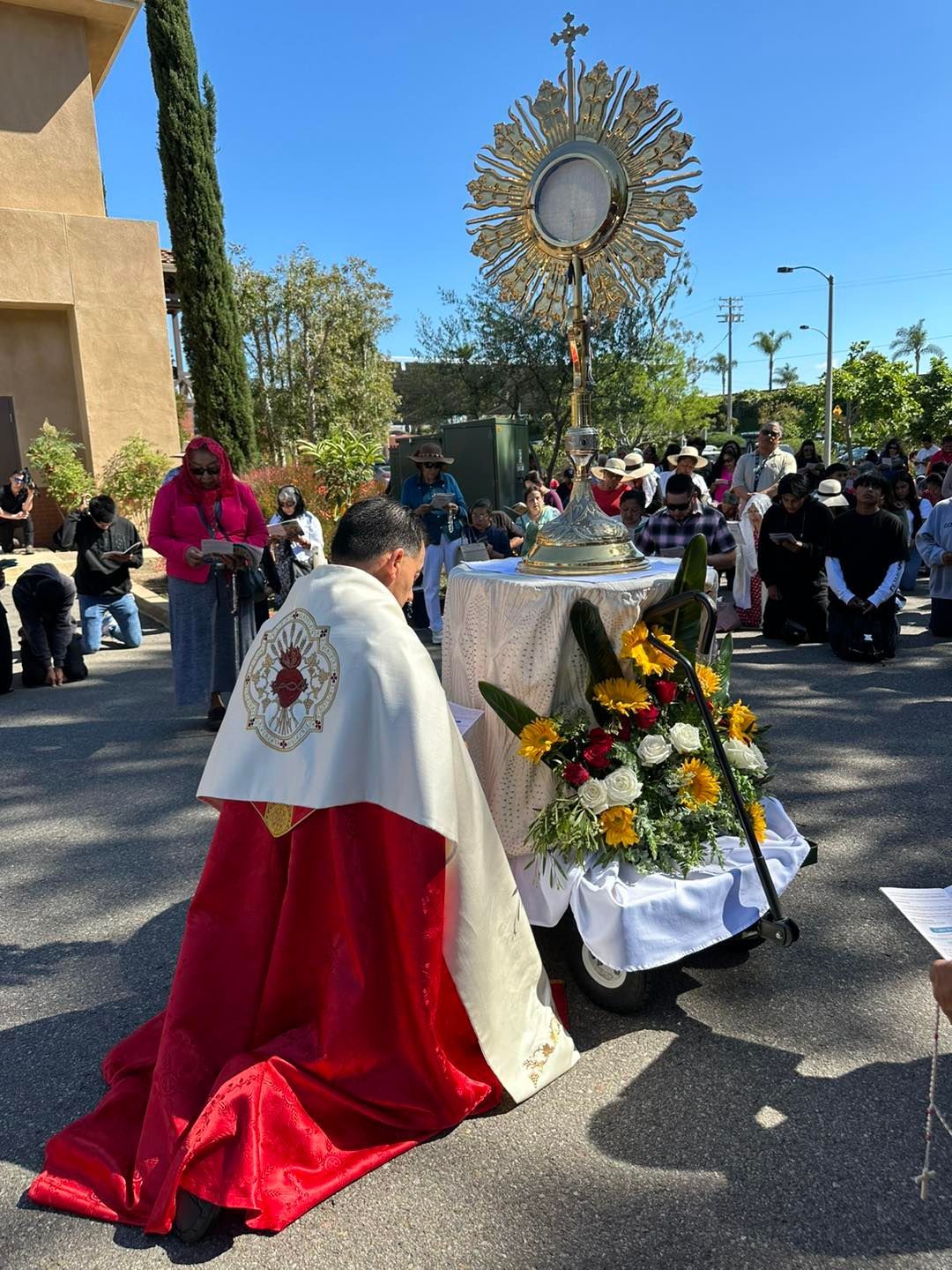 Religious procession outdoors with a priest kneeling before a flower-decorated altar and a sunburst monstrance.