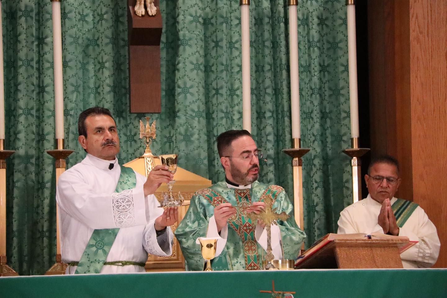 Three clergy in green vestments celebrate Mass at an altar with candles and a chalice.
