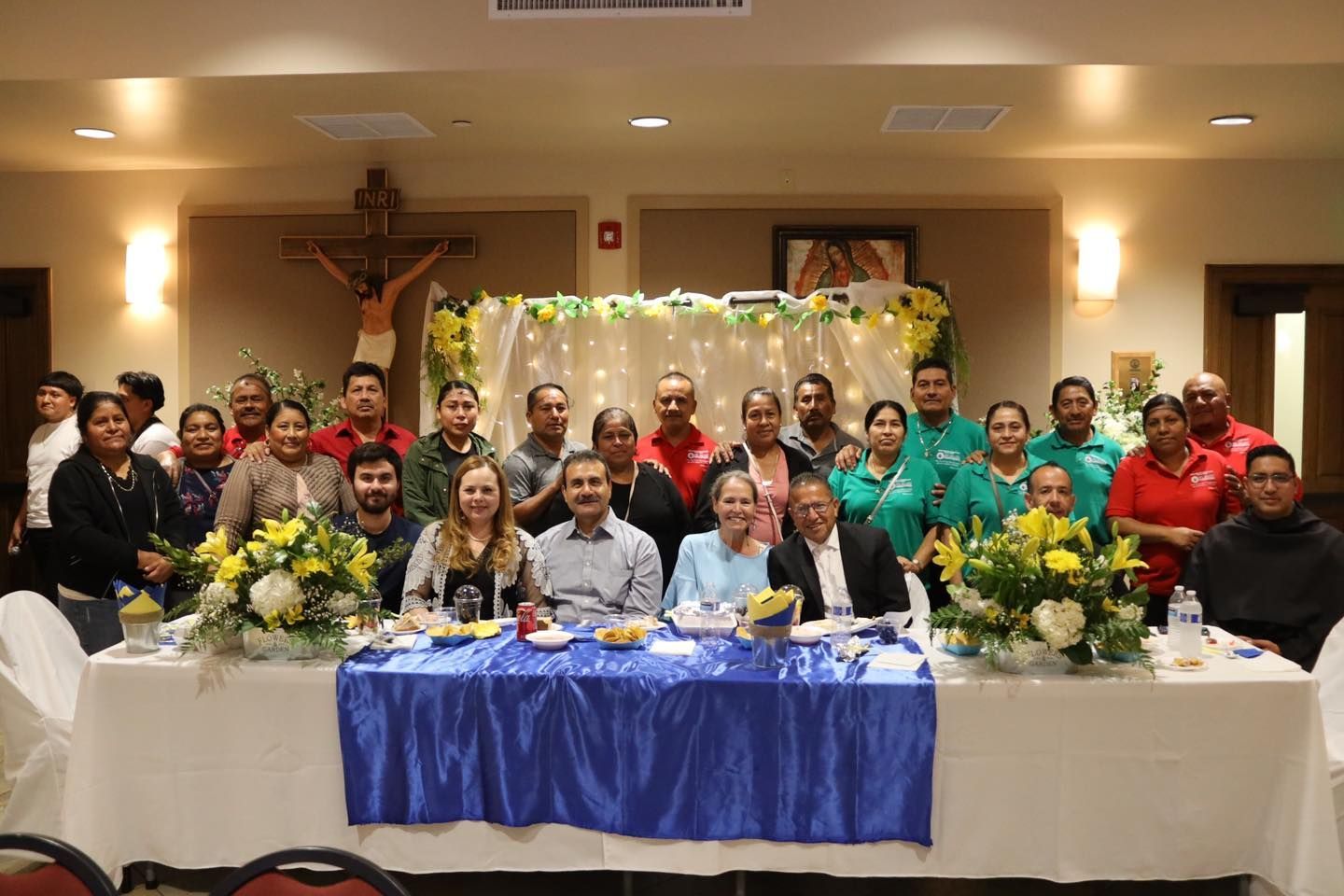 Large group posed at a banquet table with blue tablecloths in a decorated hall, flowers and a crucifix behind them