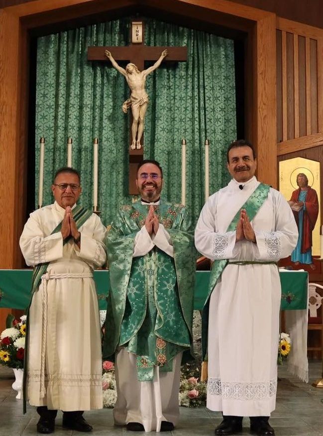 Three priests stand in prayer before an altar with a crucifix, wearing white and green vestments.