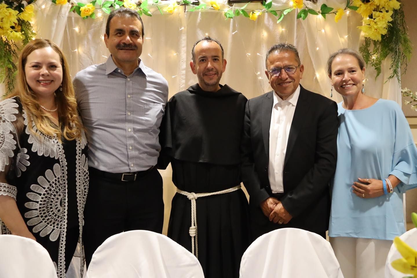 Five people posing indoors under yellow floral garlands, smiling at a formal event