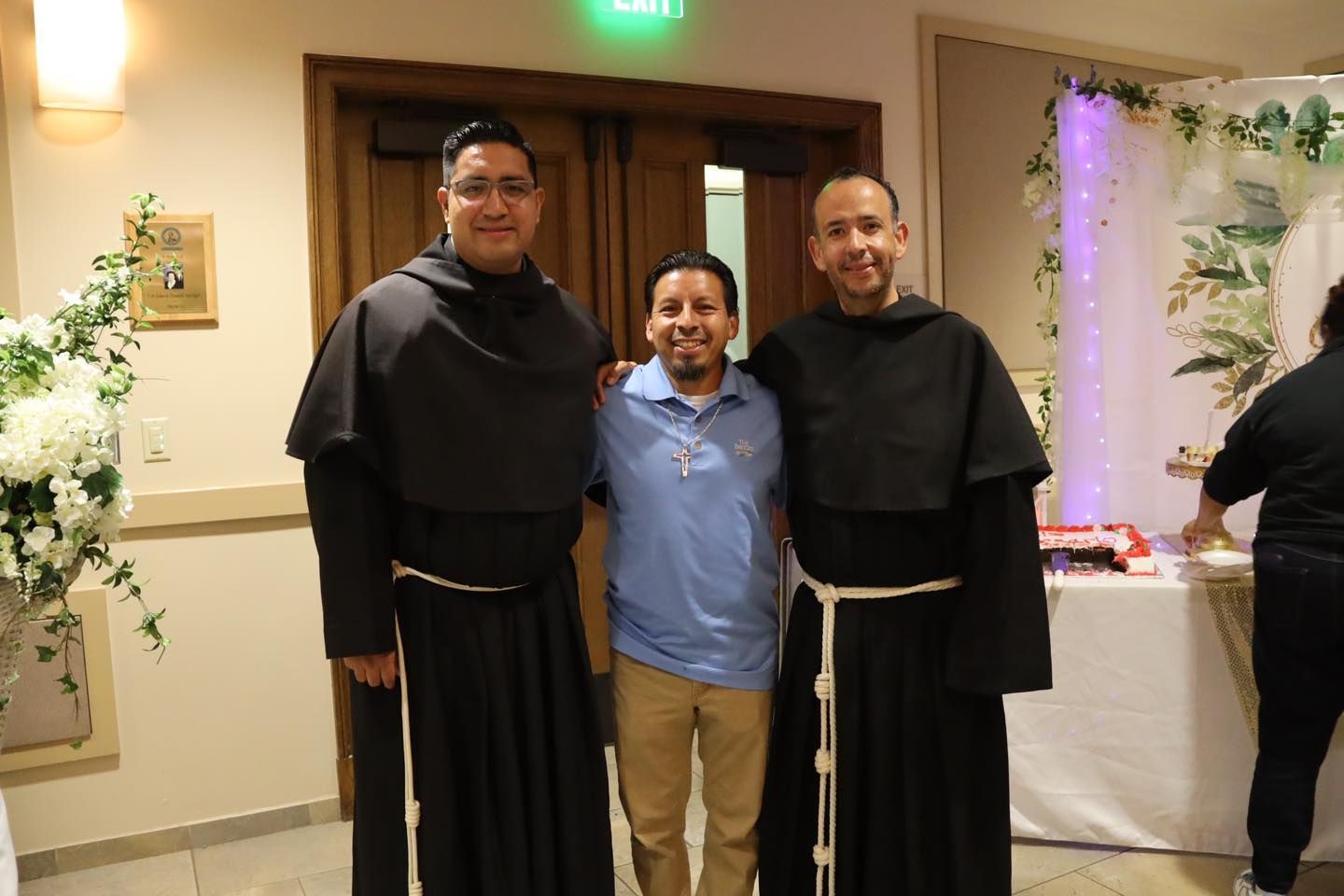 Three people standing indoors at a formal event, with two in black robes beside one in a blue shirt.